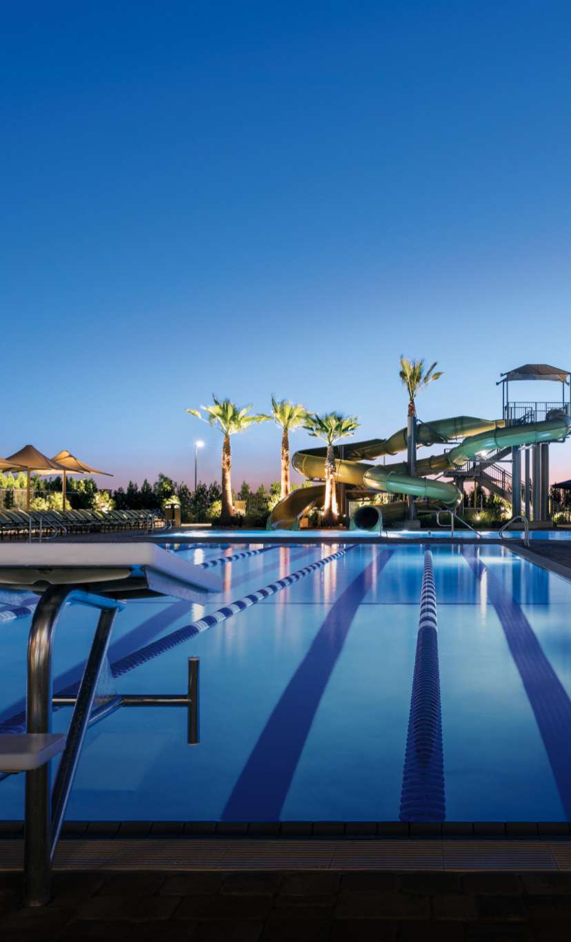 An outdoor lap pool with starting blocks and lanes, a waterslide in the distance, at dusk