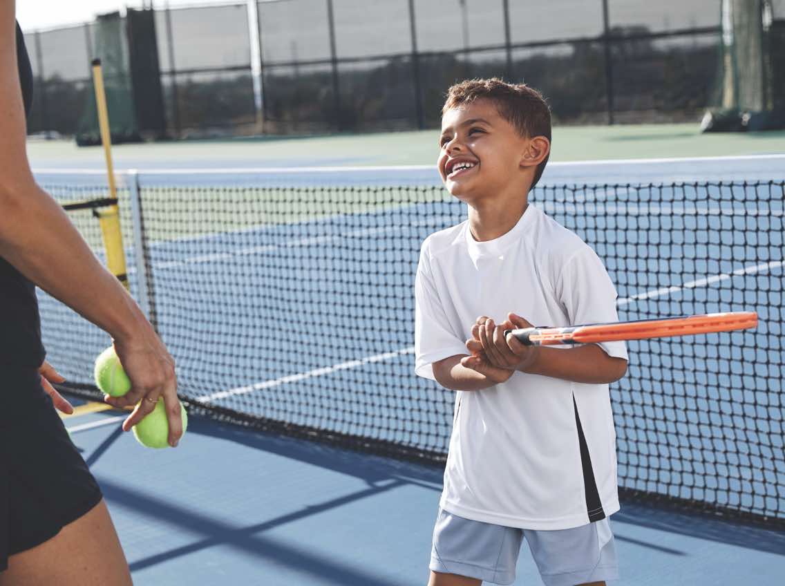 Male child smiling holding a tennis racquet on an outdoor tennis court at Life Time
