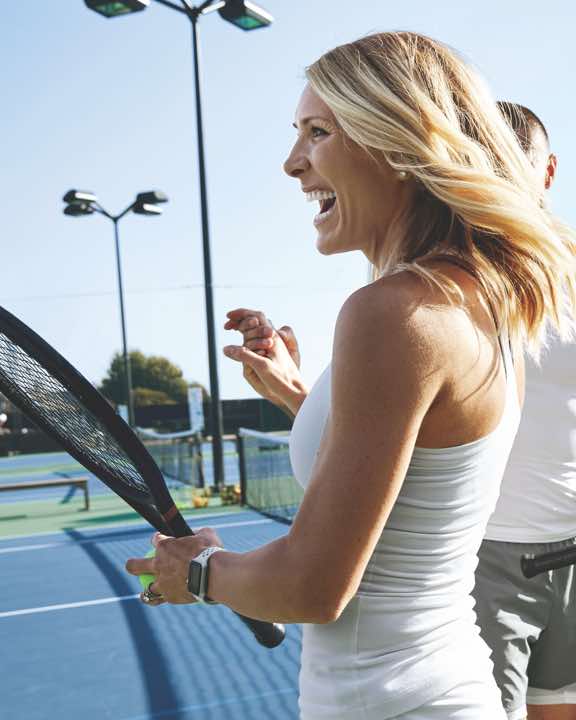 Smiling woman holding a tennis racquet on an outdoor tennis court at Life Time