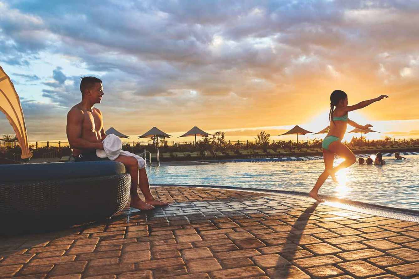 A dad watches his daughter jump into an outdoor pool at sunset