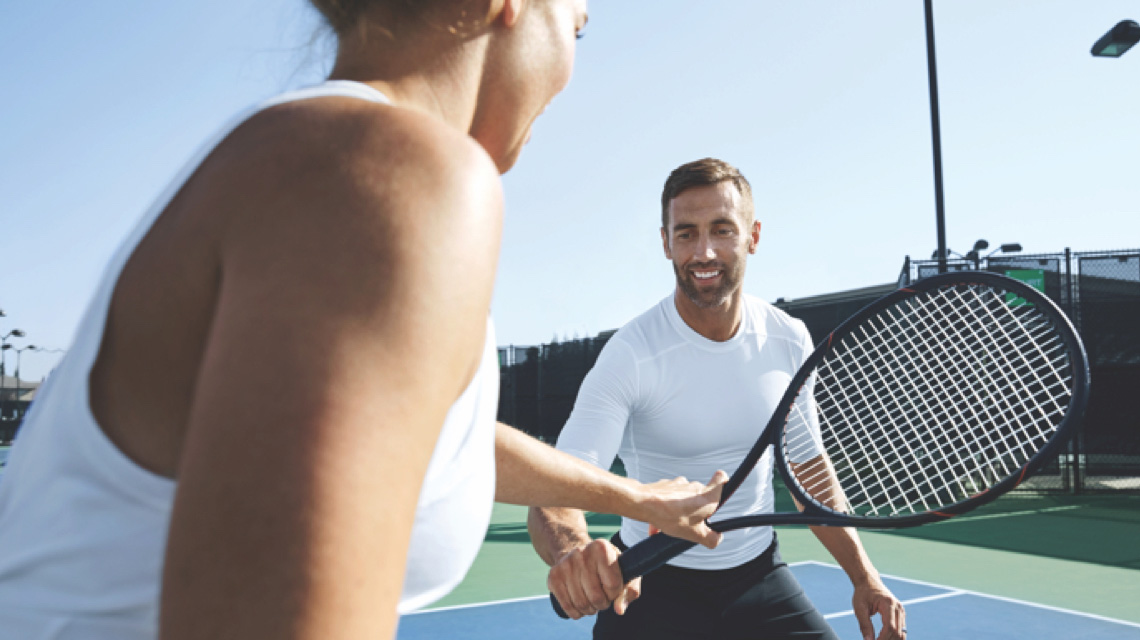 A man receives instruction on his racquet from a Life Time Tennis Instructor on an outdoor Life Time Tennis court
