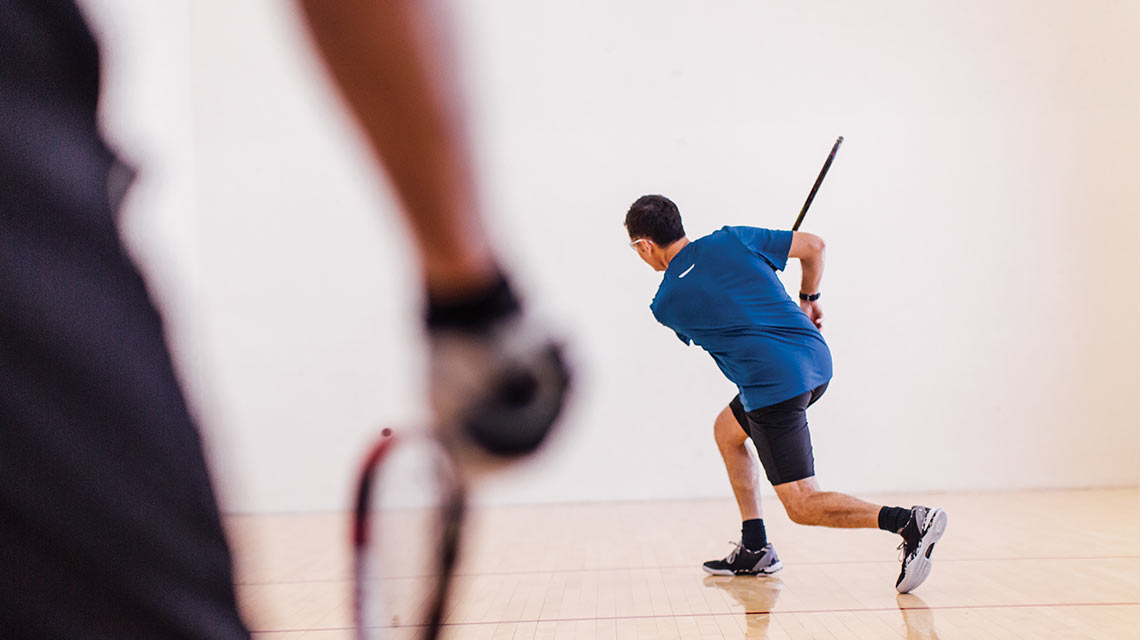 A man swings his racquet with his opponent in the foreground of the racquetball court