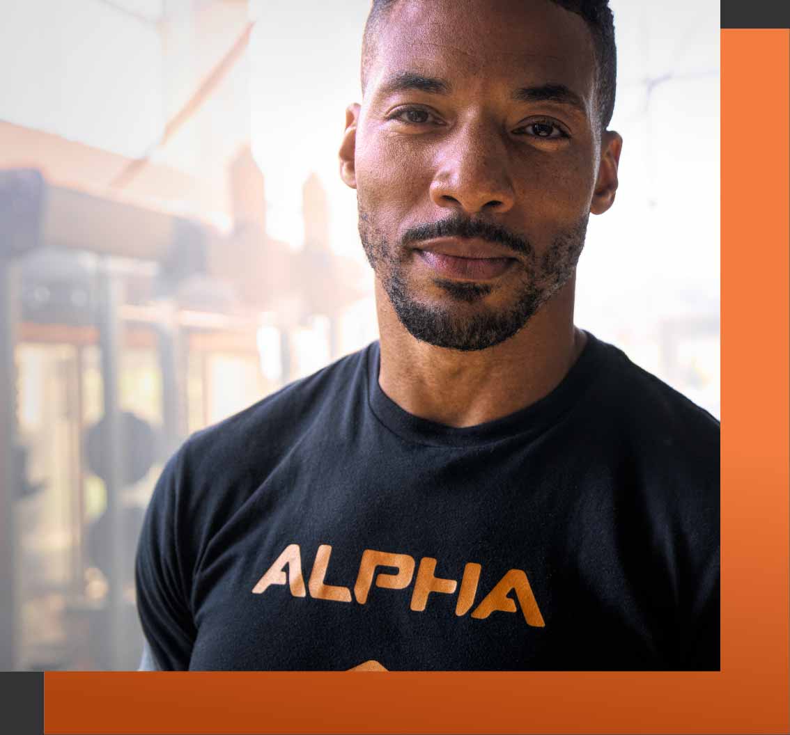 A shot of Coach Freeman wearing an Alpha shirt, weightlifting equipment in the background