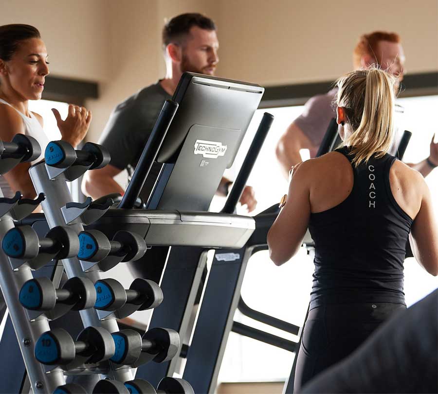 Three GTX members run on treadmills in a studio filled with beautiful morning light, a weight rack in the foreground. 