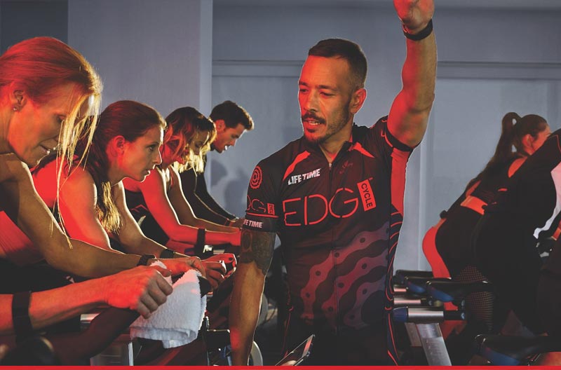 A group people cycling in a row with an instructor nearby, looking on and with an arm raised in joy. 